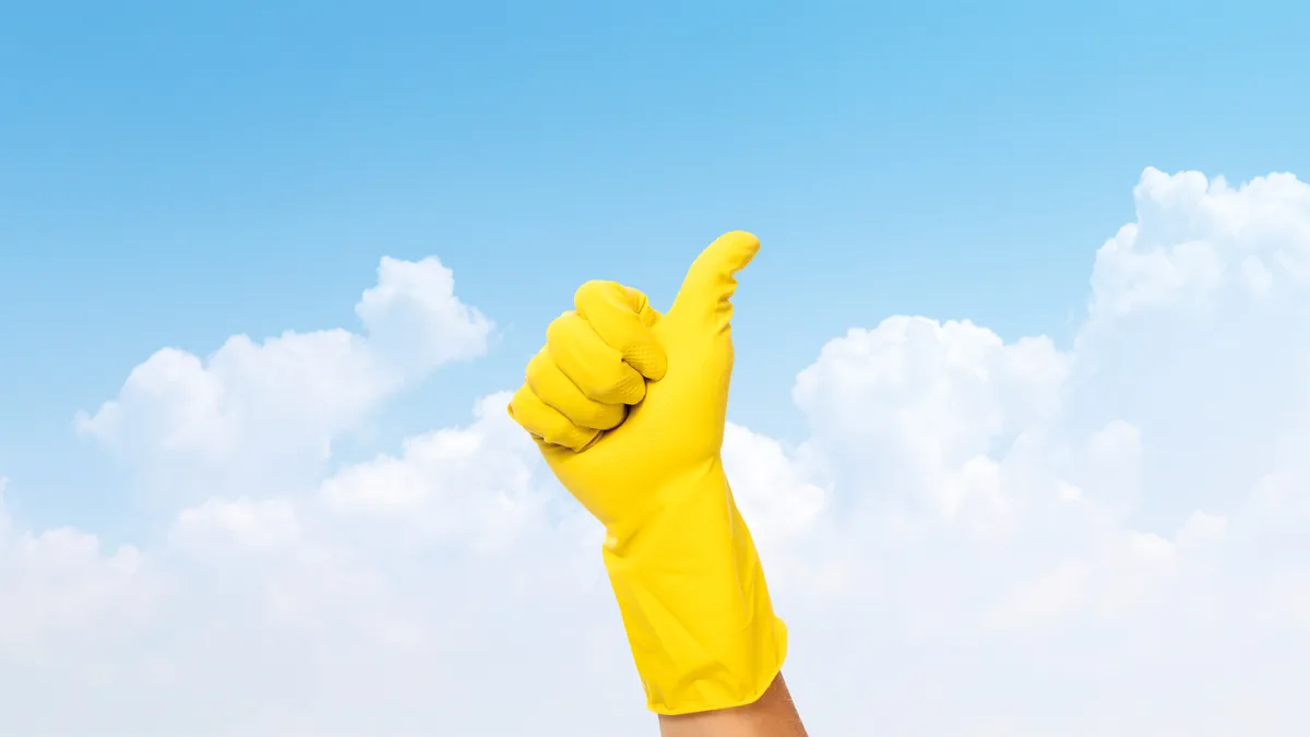 Gelber Gummihandschuh mit erhobenem Daumen vor blauem Himmel mit Wolken